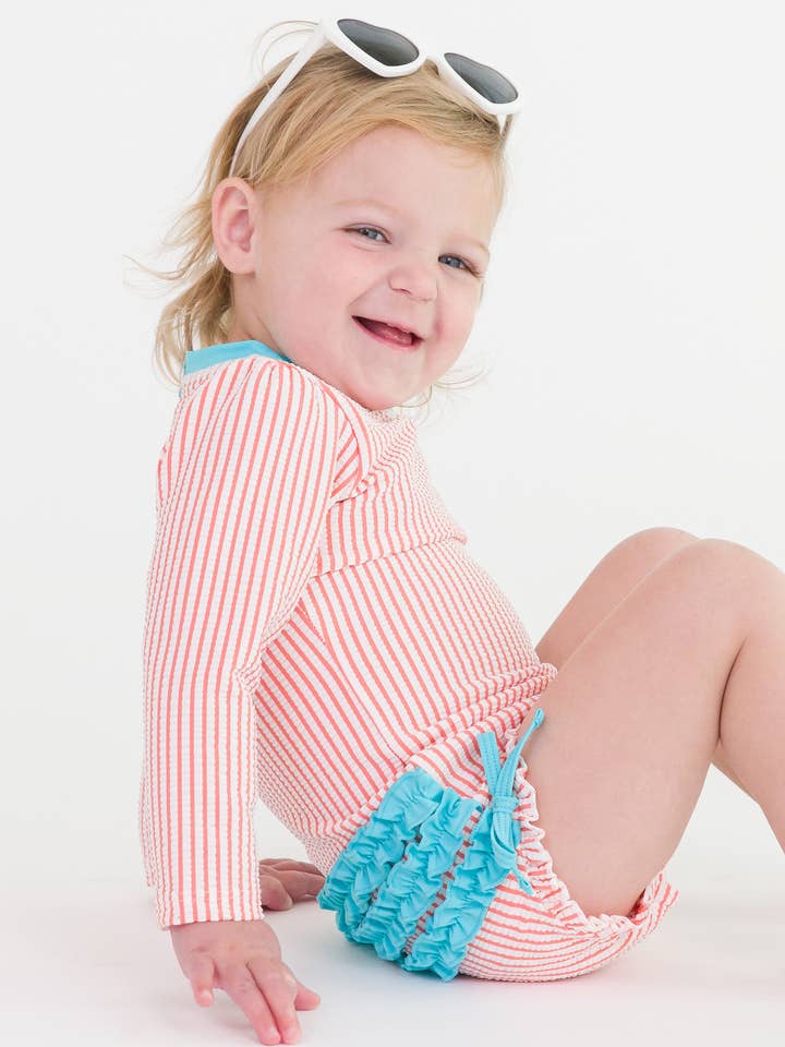 Costume intero con protezione solare a maniche lunghe in seersucker corallo per bambine con volant posteriori distintivi. per la vendita all'ingrosso da parte di RuffleButts + RuggedButts