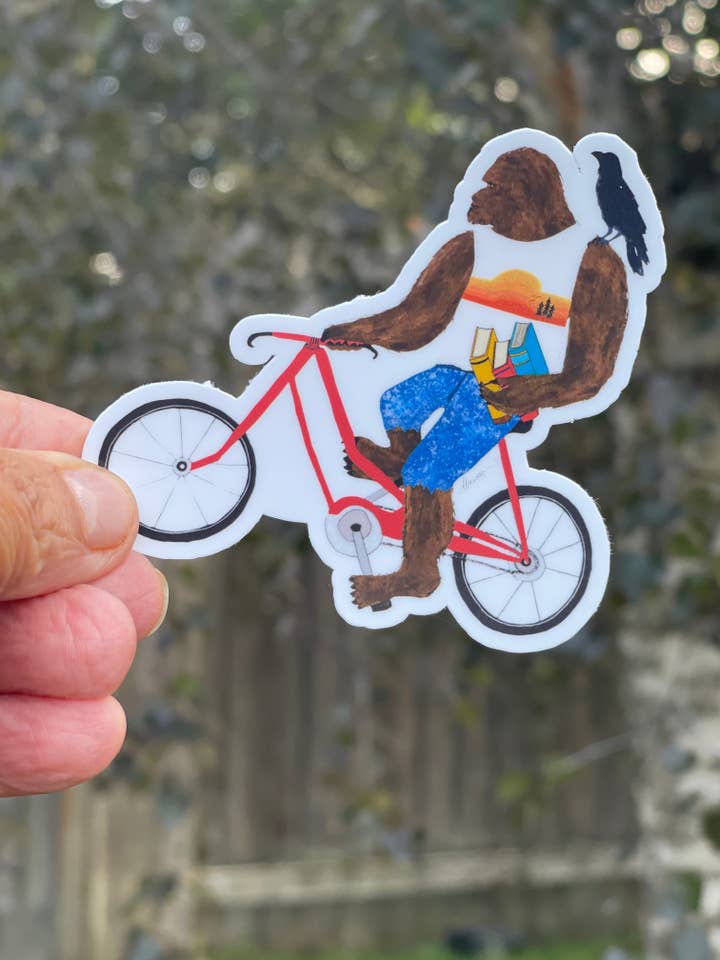 Bigfoot on Bike Sticker with Books and Crow – Whimsical Sasquatch Vinyl Decal Die Cut Sticker for wholesale by Color Crumbs Club