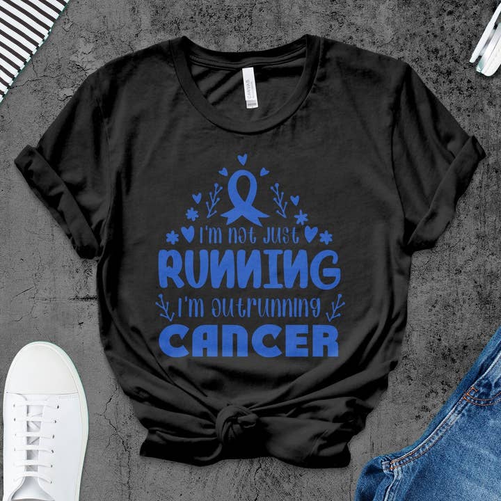T-shirt til kræftbevidsthed med 'Outrunning Cancer' motiverende citat og støttebånd for engroshandel hos FreakyTeeFactory