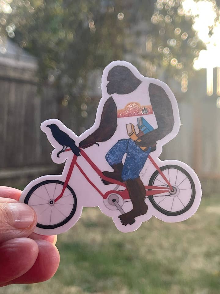 Bigfoot with books on bike with Crow – Whimsical Sasquatch Vinyl Decal Die Cut Sticker for wholesale by Color Crumbs Club