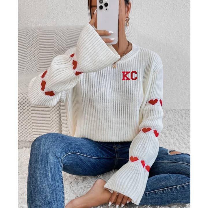Vit stickad tröja med KC bokstäver och hjärtan på ärmarna för wholesale av Elby Designs Apparel
